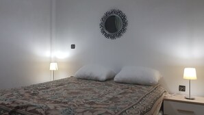 Uma cama bem arrumada com colcha estampada, duas luminárias de cabeceira, um espelho fixado na parede e uma mesinha de cabeceira com abajur.