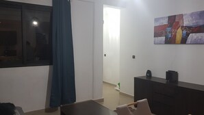 Uma sala de estar com um sofá de cor escura, uma mesa pequena, um móvel escuro com dois alto-falantes e um quadro abstrato colorido na parede.