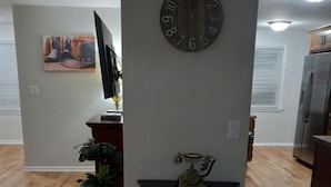 A living room with a wooden floor, a round wall clock, a flat-screen TV, a plant, and a small table with a decorative item.