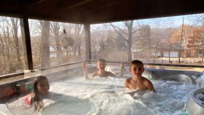 Ein Whirlpool mit drei Personen darin, umgeben von einer Holzterrasse und Blick auf Bäume sowie ein Gewässer.