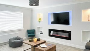 Una sala de estar moderna con televisor de pantalla plana, chimenea, un sillón color turquesa, mesa de centro de madera y una lámpara colgante.