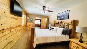 A bedroom with a wooden wall, a bed with a headboard, a nightstand, a dresser, a ceiling fan, and a framed picture on the wall.