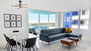 Un salon moderne avec un canapé d’angle, une table à manger avec des chaises, une table basse et un lustre. De grandes fenêtres offrent une vue sur la ville.