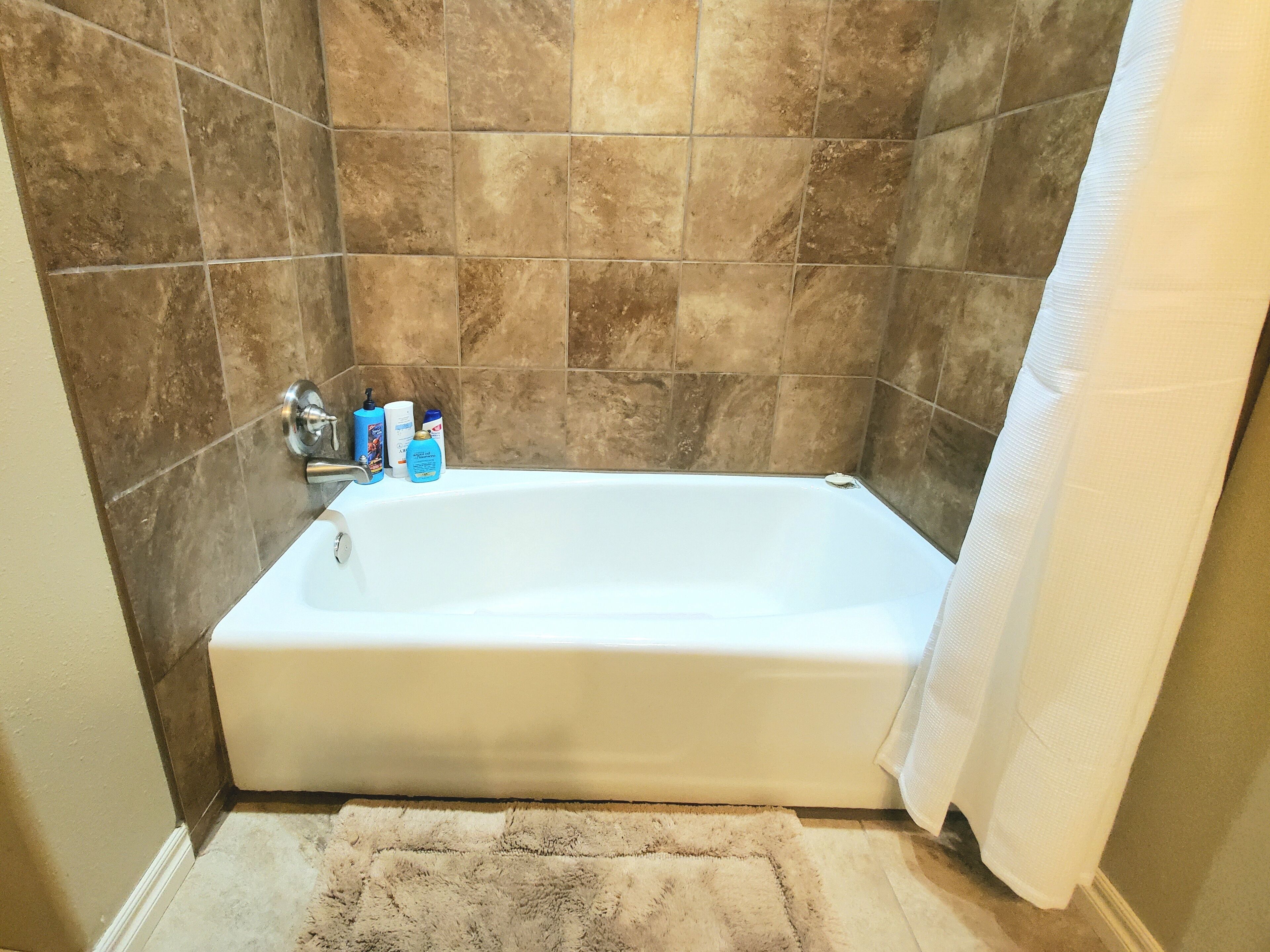 Ein Badezimmer mit einer Badewanne, braunen Fliesen, einem weißen Duschvorhang und Pflegeprodukten am Rand.
