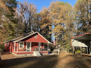 A red building with a porch, a covered area, and trees in the background. - Little Red Lake House (Greers Ferry)