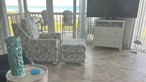 A living room with a black leather sofa, patterned ottoman, and a TV on a white cabinet. A wooden table with a decorative vase is in the foreground. The room has a view of the ocean through large windows. - 3 Bed/3 Bath ~ 2nd Floor Oceanfront Prime Condo ~ Elevator ~ 1205 Queens Court (Emerald Isle)