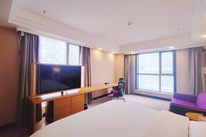 Habitación de hotel con una cama grande, un escritorio con televisión de pantalla plana, una silla, un sofá y ventanas amplias con vista a la ciudad.
