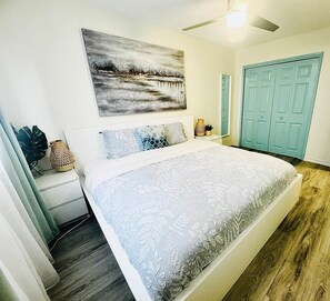 A bedroom with a bed, a ceiling fan, a wall-mounted painting, and a door.