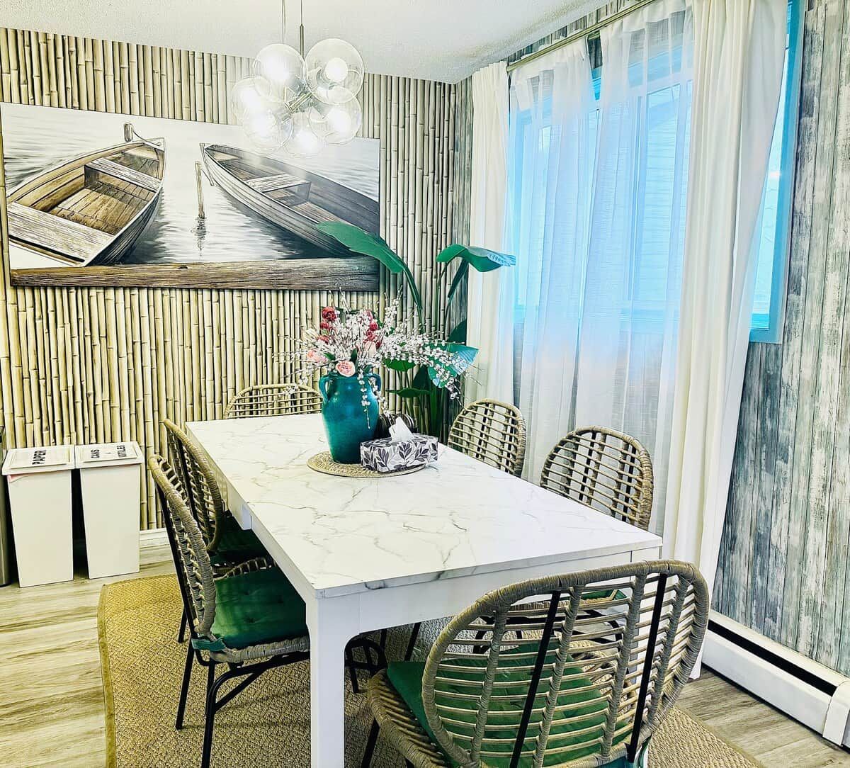Un comedor con una mesa blanca, sillas de mimbre y un cuadro de barcos en la pared.