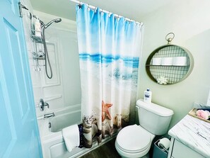 Een badkamer met een douchegordijn waarop een strandafbeelding staat, een toilet, een wastafel met spiegel en een handdoekhouder.