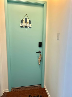 Une porte de couleur turquoise avec une pancarte indiquant « BIENVENUE », un lecteur de cartes magnétiques et un paillasson.