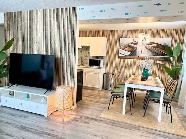 A modern living room with a flat-screen TV, a dining table with chairs, and a kitchen in the background.