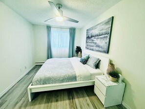 Een slaapkamer met een bed, een nachtkastje, een plafondventilator, een raam met gordijnen en een schilderij aan de muur.