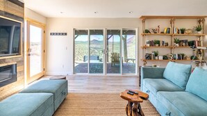 A spacious living room with a large sliding glass door leading to an outdoor patio, a built-in TV, a blue sofa, and a wooden bookshelf. - The Invisible Wind Cabin- Joshua Tree Amazing View (Yucca Valley)