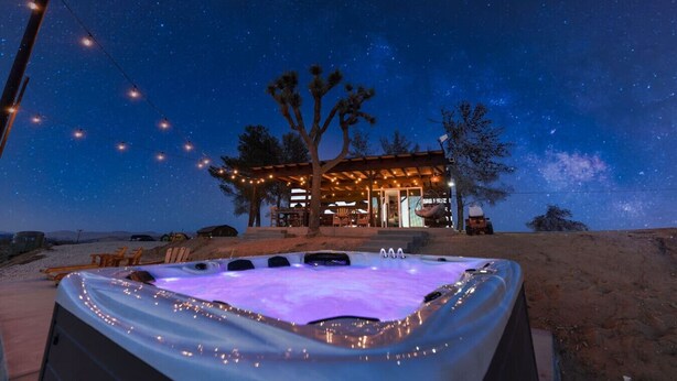 A hot tub with string lights, a wooden deck, and a building with string lights in the background. - The Invisible Wind Cabin- Joshua Tree Amazing View (Yucca Valley)
