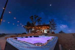 A hot tub with string lights, a wooden deck, and a building with string lights in the background. - The Invisible Wind Cabin- Joshua Tree Amazing View (Yucca Valley)
