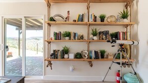 A room with a wooden bookshelf filled with books, plants, and decorative items. A telescope is set up on a tripod in front of the bookshelf. - The Invisible Wind Cabin- Joshua Tree Amazing View (Yucca Valley)