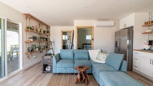 A modern living room with a sectional sofa, wooden shelves, a small table, and a kitchen area with a refrigerator. - The Invisible Wind Cabin- Joshua Tree Amazing View (Yucca Valley)