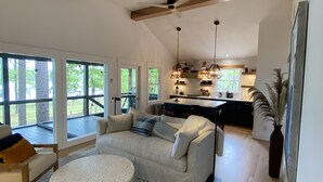 A spacious living room with a large sectional sofa, a round coffee table, and a wooden accent chair. The room features large windows, a ceiling fan, and a kitchen area visible in the background.