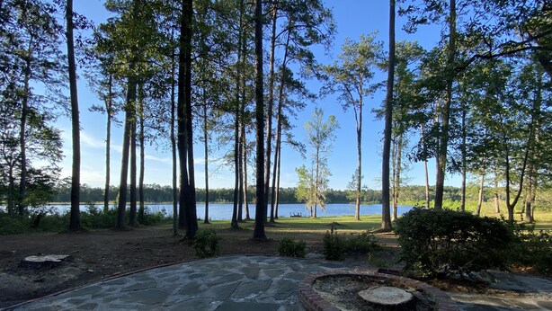 A stone patio with a circular fire pit, surrounded by a brick border, overlooking a lake with tall trees and a grassy area.