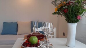 A dining table with a plate of cherries, a green apple, and two wine glasses. There is a vase with flowers and a blue pillow on a white sofa in the background. - Louis Residence (Paros)