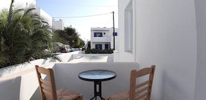A balcony with a small round table and two wooden chairs, overlooking a street with white buildings and palm trees. - Louis Residence (Paros)