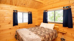 A cozy wooden cabin bedroom with a bed, two windows with curtains, a small wooden nightstand, and a trash bin.