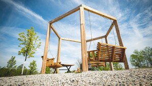 A wooden swing set with a bench, surrounded by gravel and trees, under a clear sky.
