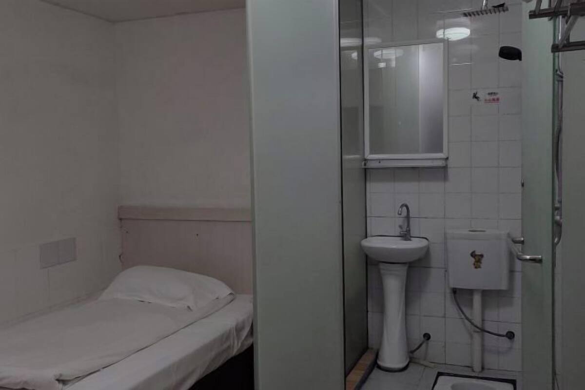 Una habitación pequeña, con poca luz, que tiene una cama individual, un lavamanos blanco y un espejo encima.