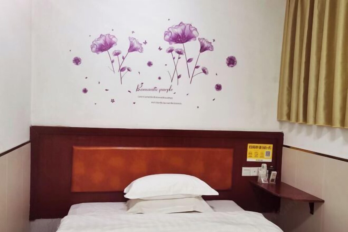 Habitación de hotel con cama, cabecera, decoración floral púrpura en la pared, cortina y una pequeña estantería con objetos.