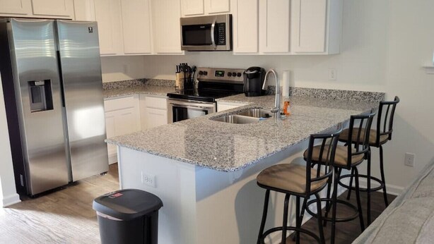 Una cocina moderna con armarios blancos, electrodomésticos de acero inoxidable, encimeras de granito y una zona con taburetes de bar.