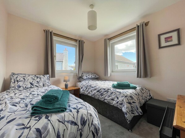 A bedroom with two beds, each with a floral-patterned duvet, a bedside table with a lamp, and a view of the outside through a window. - Cute, comfy bolthole in an amazing spot to explore from. (Kinlochbervie, near Lochinver)