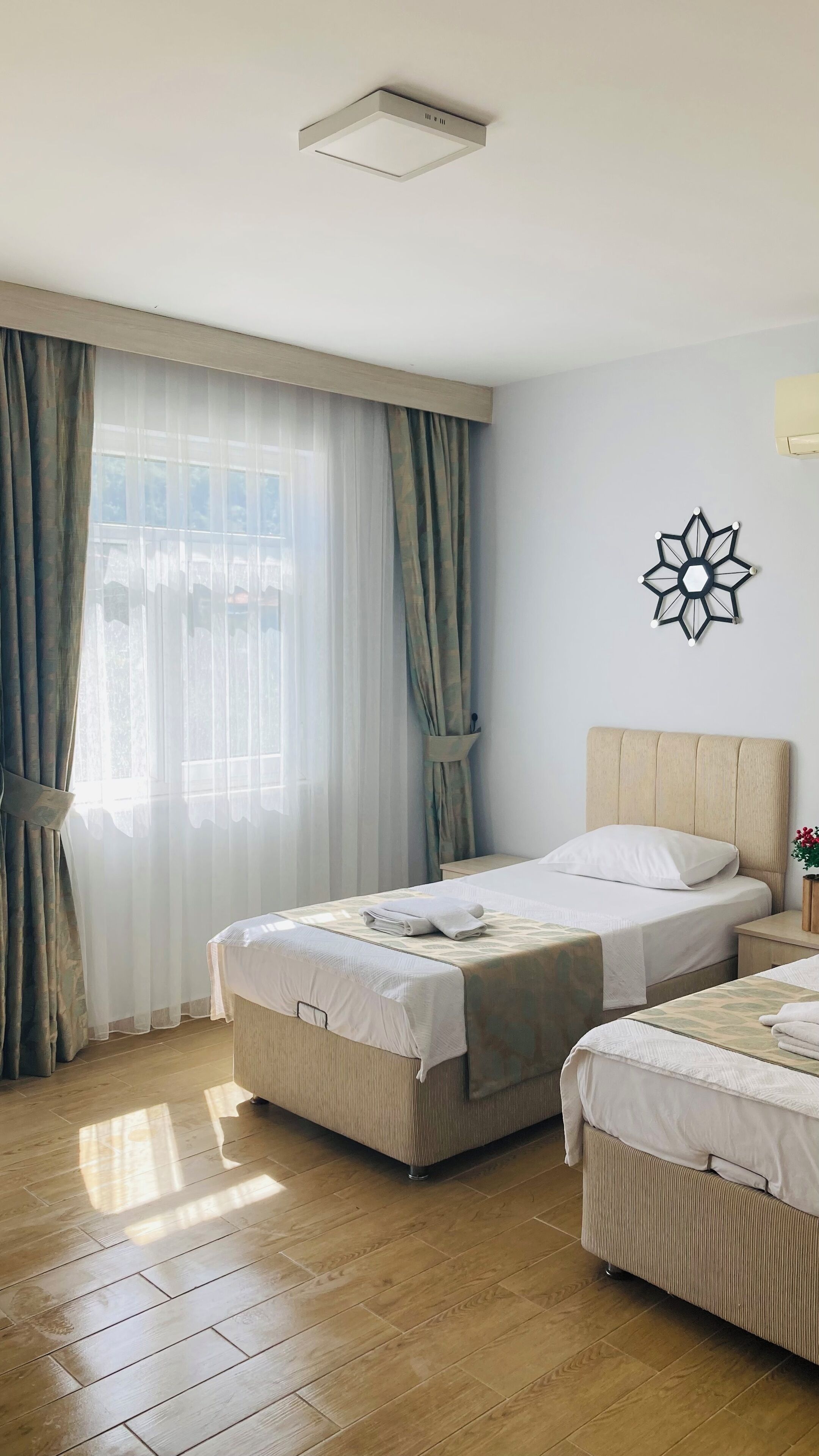 A hotel room with two beds, a large window with sheer curtains, a ceiling light, and a wall decoration.