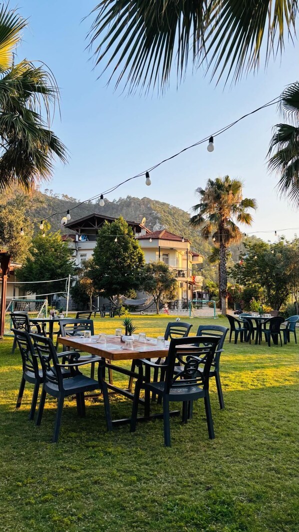Un área de comedor al aire libre con mesas y sillas, rodeada de palmeras y un edificio al fondo.