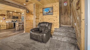 Interior de una cabaña de troncos con un sillón de cuero reclinable, paredes de madera, una zona de cocina y un cuadro de un alce.