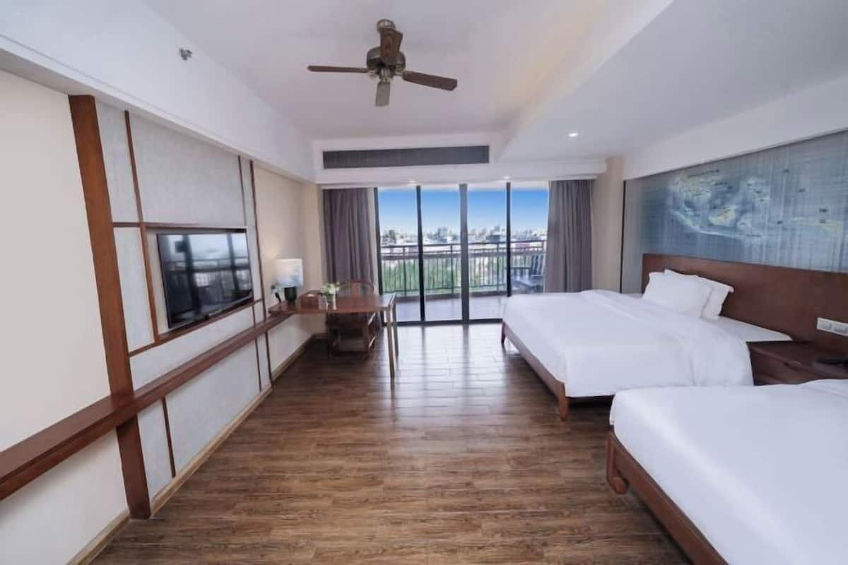 Une chambre d’hôtel avec deux lits, un ventilateur de plafond, une grande fenêtre donnant sur la ville et un plancher en bois.
