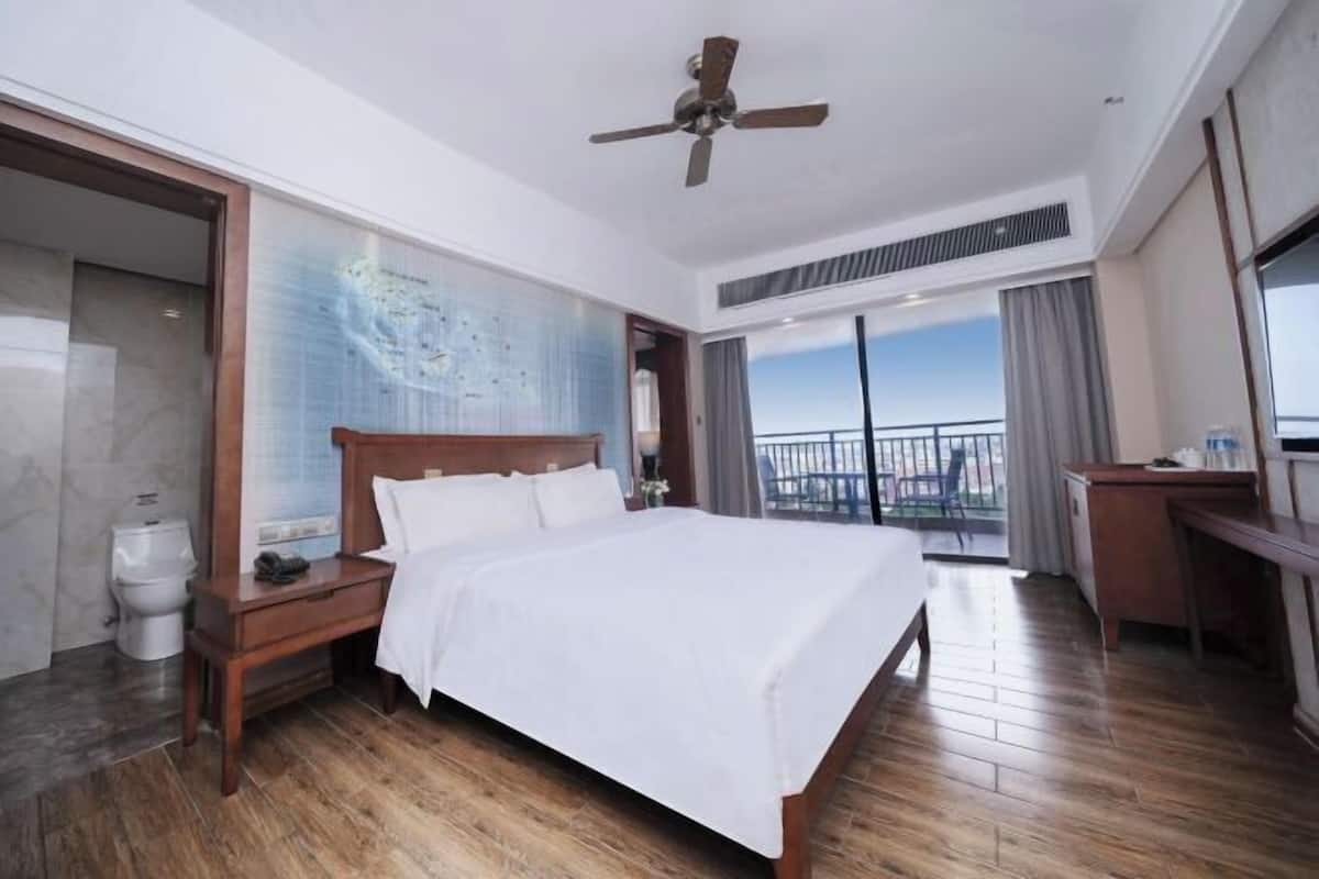Une chambre d’hôtel avec un grand lit, un plancher en bois, un ventilateur de plafond, un bureau et un balcon offrant une vue sur la ville.