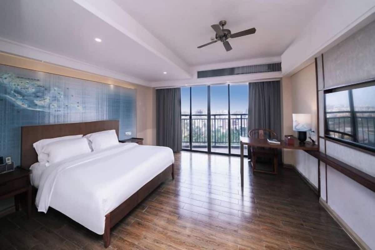 Une chambre d’hôtel avec un grand lit, un ventilateur de plafond, une vue sur la ville grâce à de grandes fenêtres et un balcon avec une chaise.