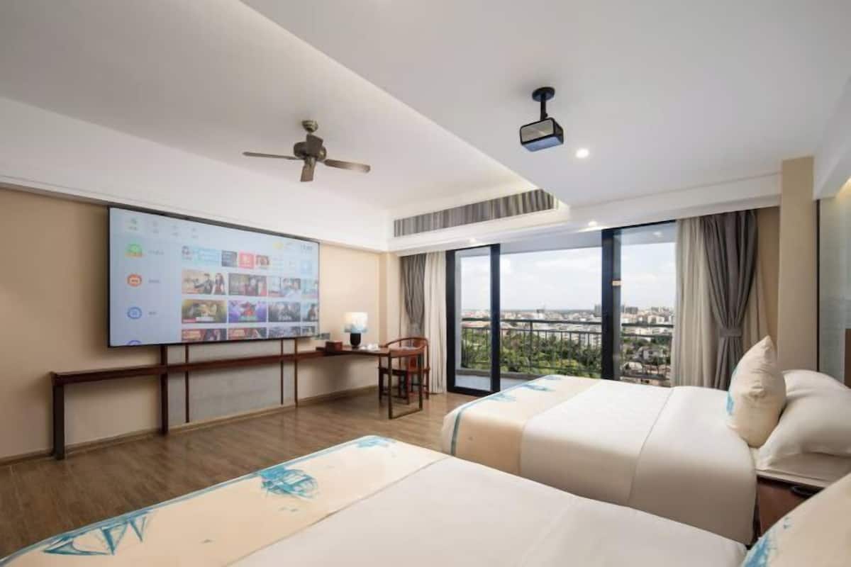 Une chambre d’hôtel avec deux lits, un bureau avec un ordinateur, une grande fenêtre donnant sur la ville et un ventilateur de plafond.