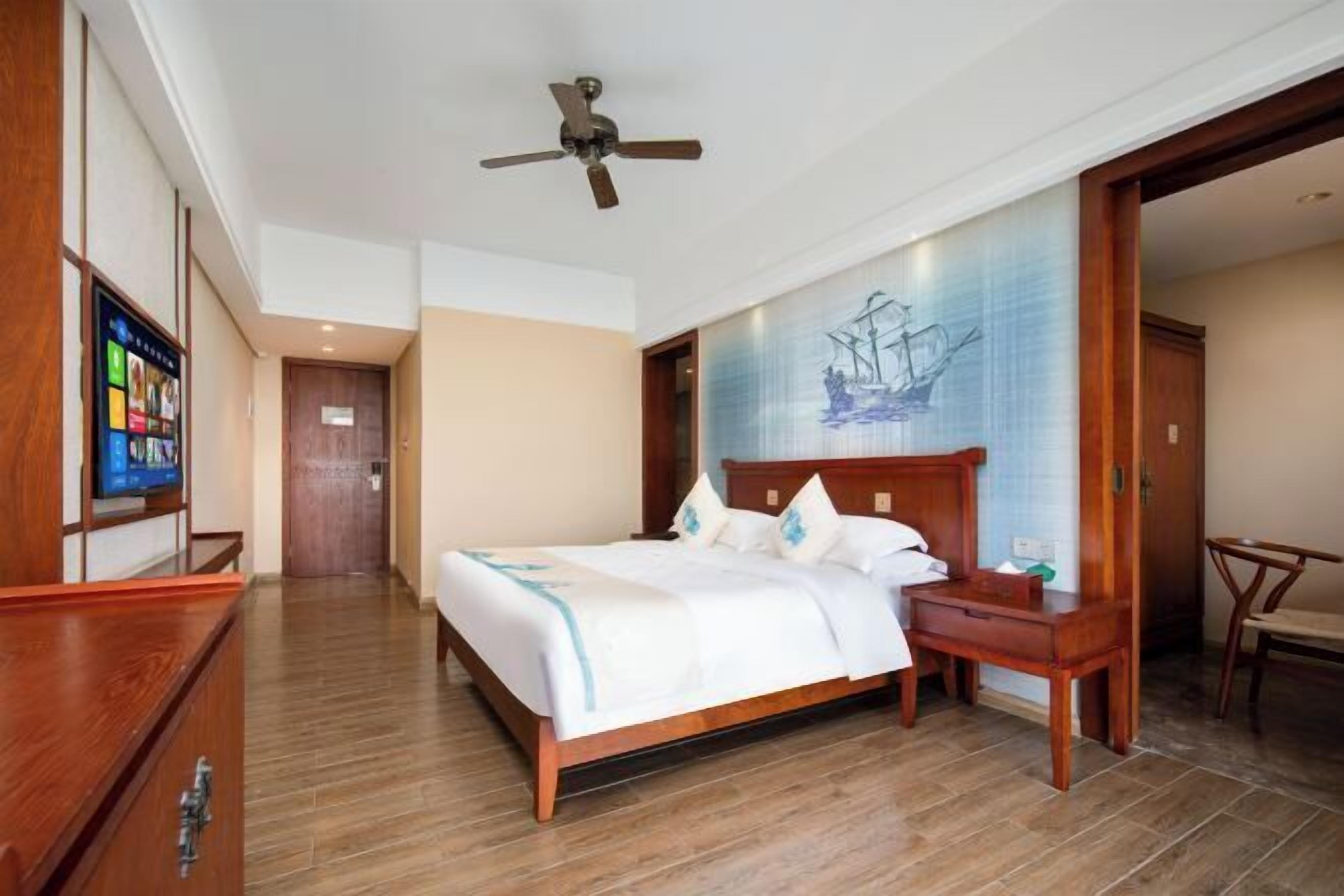 A hotel room with a wooden bed, a desk, a chair, a ceiling fan, and a wall-mounted TV.