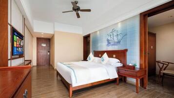 A hotel room with a wooden bed, a desk, a chair, a ceiling fan, and a wall-mounted TV.