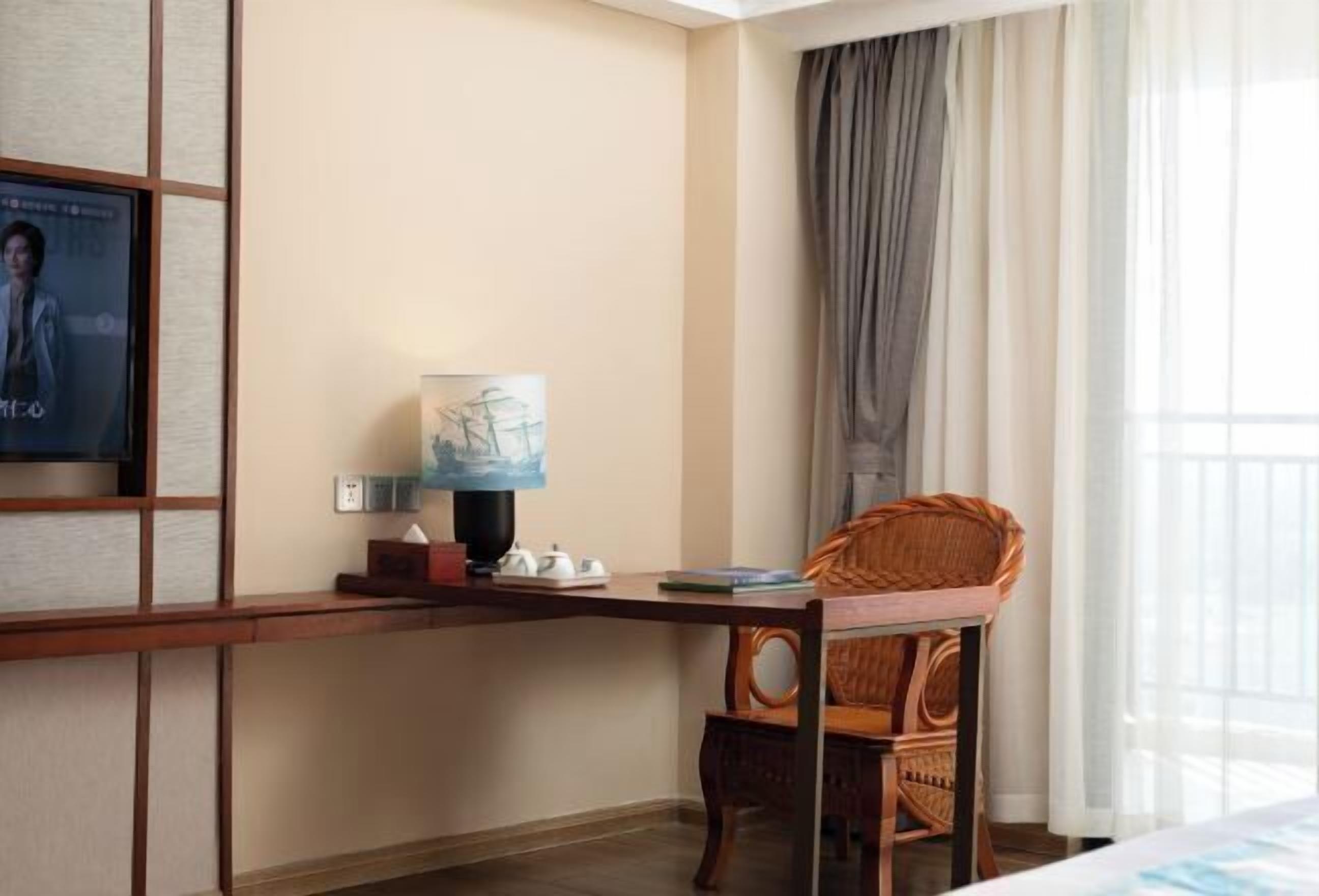 A hotel room with a wooden desk, a wicker chair, a lamp, and a poster on the wall.