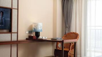 A hotel room with a wooden desk, a wicker chair, a lamp, and a poster on the wall.