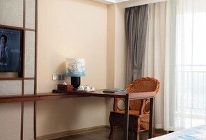 Una habitación de hotel con un escritorio de madera, una silla de mimbre, una lámpara y un cartel en la pared.