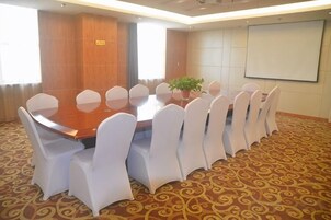 Un salón de conferencias con una gran mesa ovalada rodeada de sillas blancas, una planta en maceta y una pantalla de proyector en la pared.
