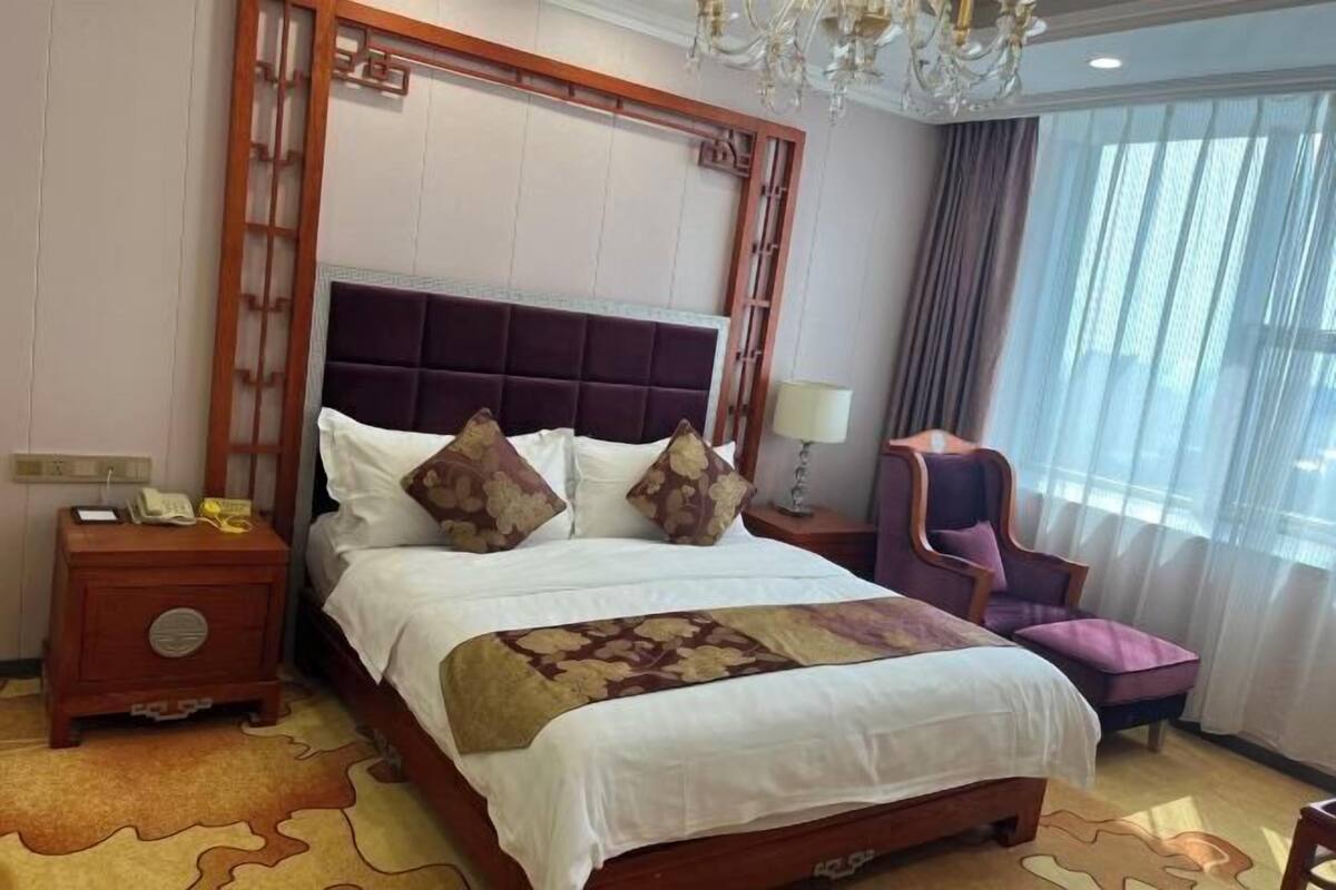 A hotel room with a large bed, a nightstand with a telephone, a chair, and a chandelier.