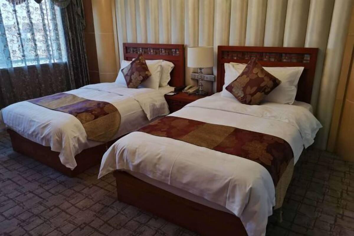 A hotel room with two beds, each with a wooden headboard, a bedside table with a lamp, and a window with curtains.