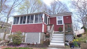 A small, single-story house with a red exterior, white trim, and a front porch with steps. - Cozy Lakeside Cottage Just Steps To The Beach (Mashpee)