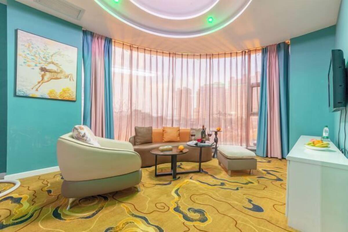Un salon moderne comprenant un canapé, un fauteuil et une table basse, un petit coin cuisine et une grande fenêtre avec des rideaux.