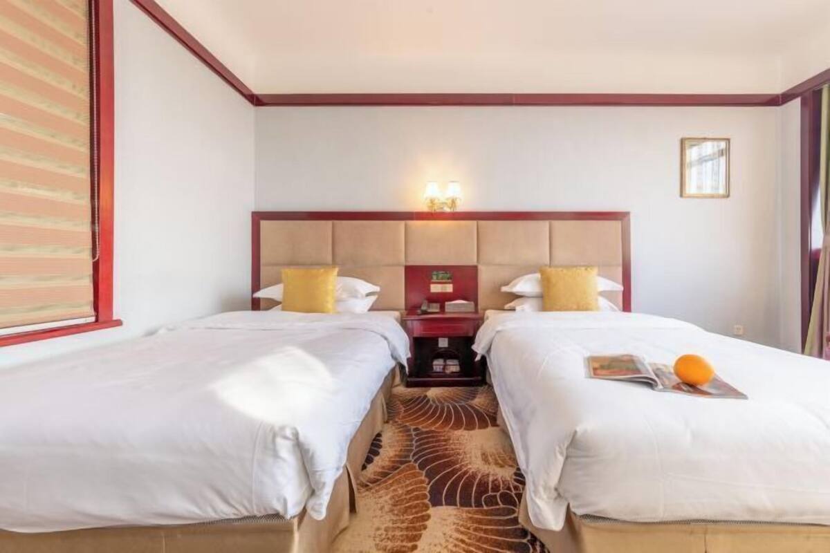 Une chambre d’hôtel avec deux lits, une tête de lit dotée d’un luminaire, une petite table et une fenêtre avec des stores.